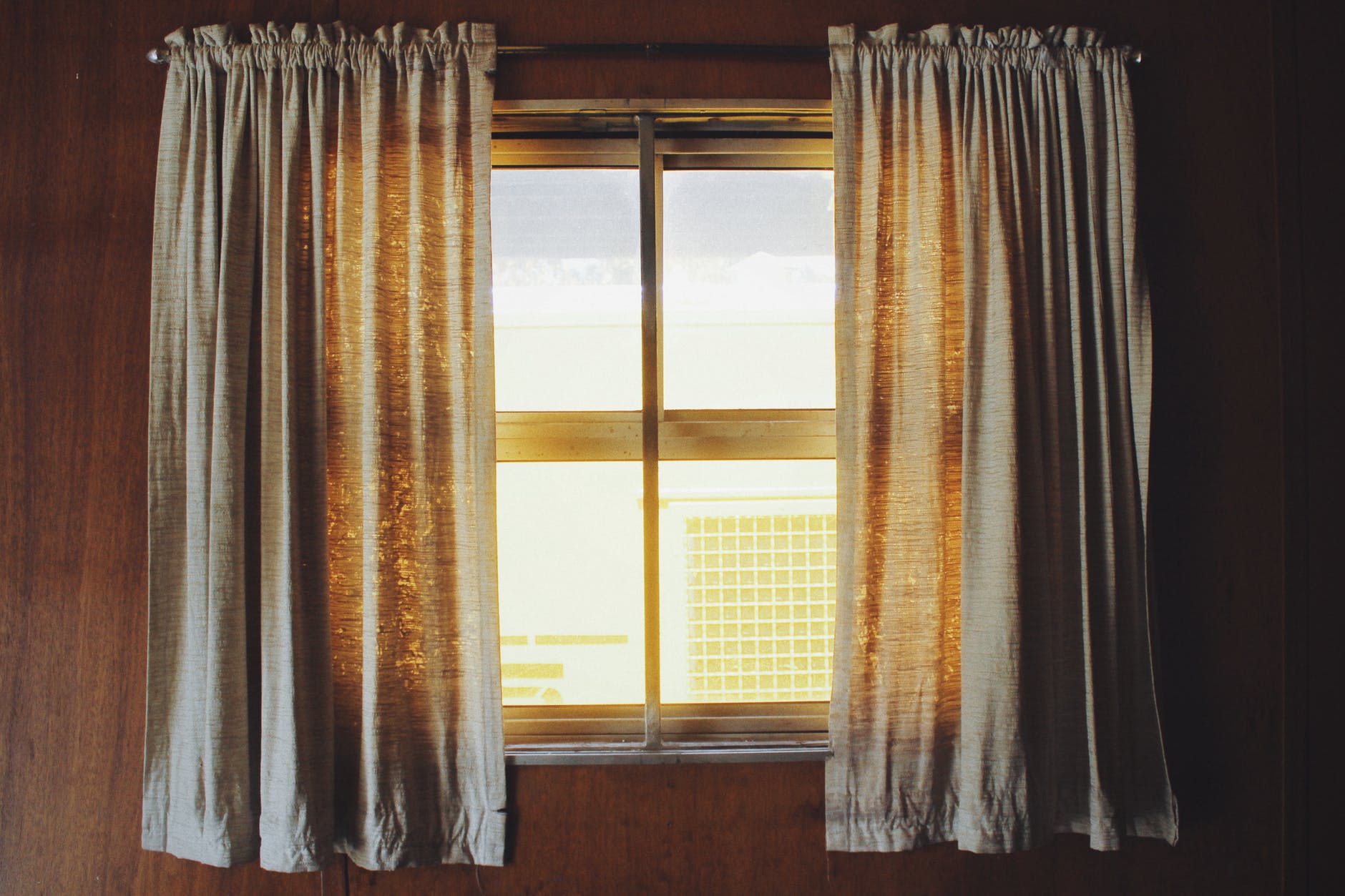 A pair of thin grey curtains hung up near a square window 