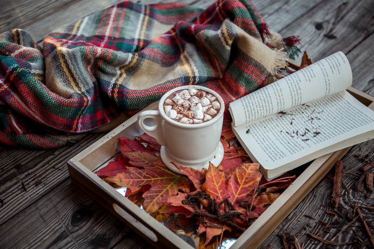 A blanket, hot drinks and book on a tray filled with autumn leaves
