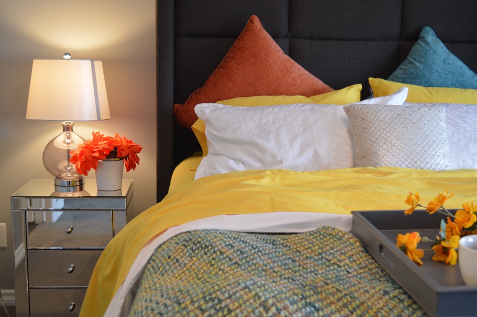 A bed with pink, yellow and blue bedding next to a bedside table with flowers.